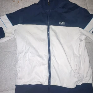 Blåvit zip hoddie från Hugo Boss - Snygg zip hoddie från Hugo Boss i marinblått och vitt med dragkedja framtill. Hoddien har ribbade muddar, två fickor och coola blå detaljer på ärmarna. Märkeslogga broderad på bröstet och grön etikett i nacken. Perfekt för dig som gillar sportig stil.