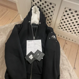 Svart Moose Knuckles hoodiejacka M - Svart hoodiejacka från Moose Knuckles med ikonisk logopatch på ärmen och dragkedja framtill. Insidan har quiltad svart foder och vit teddyfleece i huvan. Jackan har snörning i huvan och ribbade muddar. Perfekt för dig som gillar streetwear och vill ha en varm jacka med snygga detaljer. Som ni ser har jag kvitto och den är köpt ifrån miinto teddyfleecen är avtagbar.
