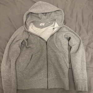 Grå hoodie från Jack & Jones S/XS - En stilren grå hoodie från Jack & Jones i storlek XS men känns mer som S. Tröjan har dragkedja framtill, huva med snörning och två fickor. Tillverkad i mjuk bomullsblandning, perfekt för chill dagar eller när du vill ha en enkel och clean look. Kan tänka mig att gå ner i pris vid snabbaffär!