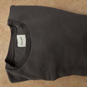 Mörkgrå sweatshirt från Jack & Jones - En stilren mörkgrå sweatshirt från Jack & Jones med rund hals och ribbade muddar. Tröjan är enkel och passar perfekt till jeans eller joggers. Materialet är mjukt och skönt bomull, och modellen har lång ärm för extra komfort.