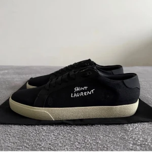 Svarta Saint Laurent sneakers strl 44 - Svarta sneakers från Saint Laurent med vit kontrastsula och handskriven logga på sidan. Klassisk låg modell med rund tå och snörning. Tillverkade i Italien med canvas och skinn för en lyxig känsla. Perfekta för dig som gillar stilrena och exklusiva sneakers.