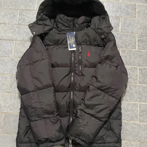 Svart pufferjacka från Polo Ralph Lauren - Svart pufferjacka från Polo Ralph Lauren med huva och flera dragkedjefickor. Jackan har en röd Polo-logga på bröstet och quiltad design. Perfekt för kalla dagar med sin tjocka och fluffiga look. Stängs med dragkedja och har justerbar huva.