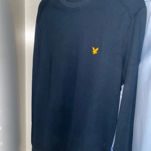 Mörkblå långärmad tröja Lyle & Scott - Snygg mörkblå långärmad tröja från Lyle & Scott med klassisk gul fågel-logga på bröstet. Tröjan har rund hals och är gjord i mjuk bomull som är skön mot huden. Passformen är normal och den funkar perfekt till jeans eller chinos.