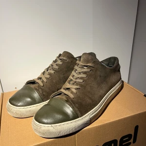 Bruna och gröna sneakers från AXEL ARIGATO i mocka - Snygga bruna och gröna sneakers från Axel Arigato  i mocka med olivgröna tåpartier och vita gummisulor. Har använt dom ett tag och det finns täcken på användning. Skorna har klassisk snörning och låg profil, perfekt för en clean och stilren look. Materialet är mjukt och ger en exklusiv känsla.