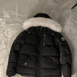 HELT NY Moose Knuckles jacka 3Q  - Hör av dig innan! Svart dunjacka från Moose Knuckles med vit fuskpäls på huvan, flera dragkedjor och snygga metallknappar. Jackan har broderad hockeypatch på insidan och ikonisk logga på ärmen. Perfekt för kalla dagar med sin tjocka och puffiga design.