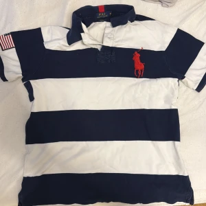Randig pikétröja från Polo Ralph Lauren - Snygg pikétröja med breda marinblå och vita ränder från Polo Ralph Lauren. Tröjan har korta ärmar, klassisk krage och stor röd logga på bröstet. Amerikansk flagga på ena ärmen ger extra stilpoäng. Perfekt för dig som gillar sportig och preppy look.