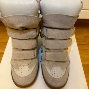 Beige sneakers Isabel Marant med kardborre - Snygga beige sneakers från Isabel Marant med högt skaft och flera breda kardborreband. Skorna har en rund tå, platt sula och är tillverkade i mocka och skinn. De har en unik, chunky design och coola paneldetaljer som ger extra edge till din stil.