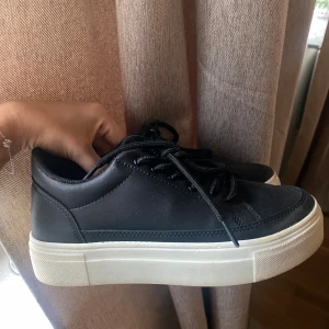 Svarta sneakers med vit platt sula - Snygga svarta sneakers från Nelly med vit platt sula och rund tå. Skorna har snörning och är gjorda i skinnimitation med stilren design. Perfekta för dig som gillar enkel och clean look. Passar till jeans, kjol eller shorts. Storlek 36, normala i storleken. Använda men i bra skick!