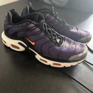 Nike Air Max Plus Tn lila sneakers - Nike Air Max Plus Tn sneakers i lila och svart med orange swoosh. Skorna har en vågig design på ovandelen, synliga luftkuddar i sulan och svarta snören. Sulan är vit med orange och gula detaljer undertill. Materialet är syntet och mesh för en sportig vibe.