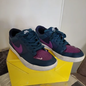 Nike SB sneakers blå/lila strl 42 - Nike SB sneakers i cool blå och lila färg med vit sula och lila swoosh. Låg modell med snörning och perforerad tå för extra ventilation. Skorna har en snygg kontrast mellan mörkblått och lila, perfekt för dig som gillar streetstyle och skate. Materialet är syntet och skinn.