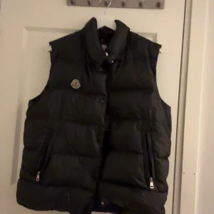 Svart dunväst från Moncler - Svart dunväst från Moncler med hög krage och quiltad design. Västen har två praktiska fickor med dragkedja och Moncler-logga på bröstet. Perfekt för lager-på-lager och håller dig varm under kyliga dagar. Pris ej hugget i sten