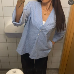 Blå randig skjorta - Blå henley skjorta med tunna vita ränder och v-ringad hals. Skjortan har tre knappar framtill och är långärmad med en loose passform. Tillverkad i mjuk bomull, perfekt för en chill och stilren look.