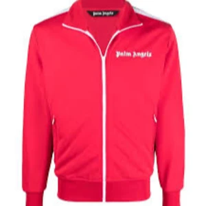 Röd tracksuit från Palm Angels - Säljer en röd tracksuit från Palm Angels med vit logga på både jacka och byxa. Jackan har dragkedja och ribbade muddar, medan byxorna har elastisk midja. Snygg och sportig look med vita detaljer längs axlarna. Perfekt för dig som gillar streetwear.
