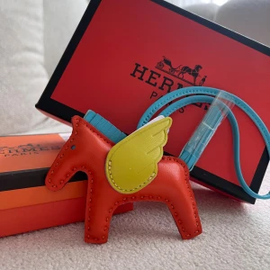  Pegasus väsktillbehör i skinn - Unik  Pegasus väsktillbehör i form av en häst med vingar. Tillverkad i orange, blått och gult skinn med fina sömmar och detaljer. Perfekt som accessoar till din väska för att ge den en färgglad och lekfull touch.