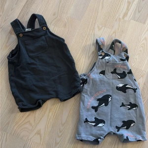 Två babyshorts H&M, svart & grå med valar - Två gulliga babyshorts från H&M i storlek 74. Ena paret är helsvart med bröstficka och knappar på axelbanden, det andra är grått med svartvita valar och texten 'WALKIN' BEACH'. Båda har korta ben och är i mjuk bomull, perfekta för varma dagar.