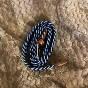 Randigt blått och vitt bälte med läderdetaljer - Säljer ett snyggt randigt bälte i blått och vitt med bruna läderdetaljer vid spännet. Bältet har ett klassiskt metallspänne och är gjort i ett flätat syntetmaterial som ger en cool look. Perfekt för att lyfta din outfit med lite extra färg och stil.