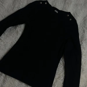 Svart ribbad stickad tröja H&M - Svart ribbad stickad tröja från H&M med snygga silvriga knappar på axlarna. Tröjan har lång ärm. 