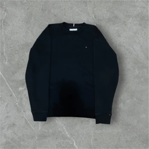 Svart sweatshirt från Tommy Hilfiger - Svart sweatshirt från Tommy Hilfiger med diskret logga på bröstet och klassisk ribbad krage. Tröjan har långärmade muddar och är gjord i mjuk bomull, perfekt för chill dagar. Snygg detalj med röd och vit rand på insidan av kragen.