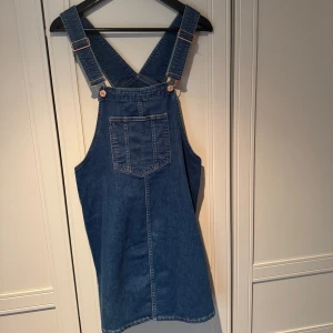 Jeansklänning med hängslen MAMA - Snygg jeansklänning från MAMA i storlek S. Modellen har breda hängslen, en stor ficka framtill och coola knappar längs sidorna. Klänningen är kort och har en klassisk blå denimlook som passar perfekt till sneakers eller boots. Använd vid fåtal tillfällen under graviditet.