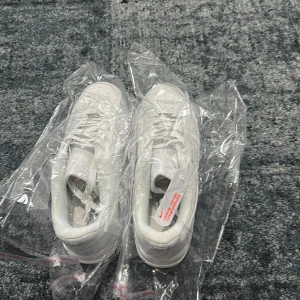 Nike Air Force 1 helvita sneakers - Nike Air Force 1 sneakers i helvit färg med klassisk låg siluett. Skorna har snörning, perforerad tå och en platt, mönstrad sula. Tillverkade i skinn med mjukt foder och tydlig Nike-logga på sidan. Perfekt för dig som gillar cleana och tidlösa sneakers.