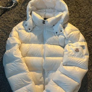 Vit Moncler pufferjacka med huva - Säljer en snygg vit pufferjacka från Moncler med huva och dragkedja framtill. Jackan har Moncler-logga på ärmen och praktisk ficka med knapp. Den är quiltad och har en riktigt clean look, perfekt för dig som gillar stilren vinteroutfit.