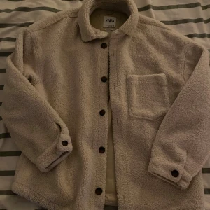 Beige teddyjacka från Zara L-XL - Säljer en beige teddyjacka från Zara i relaxed fit. Jackan har mjukt, fluffigt material, klassisk krage, knappstängning och en bröstficka. Perfekt för dig som gillar oversized och mysiga vibes. Passar dig som vill ha en chill och trendig look.
