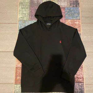 Svart hoodie - Polo Ralph Lauren - Svart hoodie från Polo Ralph Lauren med klassisk röd logobrodering på bröstet. Tröjan har huva, långa ärmar och en skön, mjuk bomullskvalitet. Perfekt för dig som gillar stilrena och tidlösa plagg med en sportig touch. Den är i storlek L men den borde varit i S, eftersom den sitter som en S. Tröjan är använd ett väldigt fåtal gånger🐦‍🔥