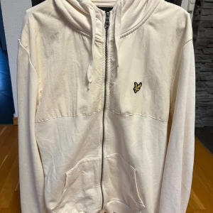 Vit hoodie från Lyle & Scott med dragkedja - Säljer en vit hoodie från Lyle & Scott med gul fågel-logga på bröstet. Tröjan har huva med snörning, praktisk dragkedja och fickor framtill. Tillverkad i mjuk bomull, perfekt för chill dagar. Långärmad och klassisk passform.