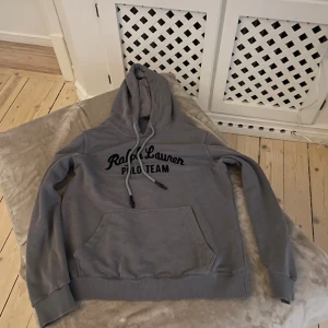 Grå hoodie från Ralph Lauren Polo - Snygg grå hoodie från polo ralph lauren med svart tryck på bröstet. Tröjan har huva med snörning, stor magficka och ribbade muddar vid ärmslut och nederkant. Perfekt för chill dagar och streetstyle. Den är Xl i barn storlek så skulle säga att den passar S.
