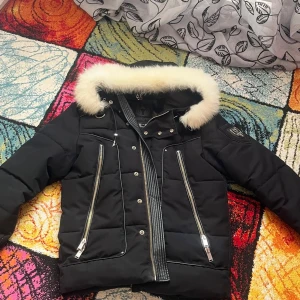 Svart Horspist vinterjacka med päls - Svart dunjacka från Horspist med fluffig vit päls runt huvan, långa ärmar och två snedställda dragkedjefickor fram. Jackan har quiltad design, silverfärgade detaljer och en snygg badge på ärmen. Perfekt för kalla dagar med stil.