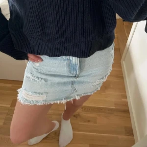 Ljusblå jeanskjol med slitningar - Säljer en ljusblå jeanskjol med rå kant och slitna detaljer framtill. Kjolen är kort och har en klassisk femficksdesign. Perfekt för dig som gillar en avslappnad och trendig stil. Passar till sneakers eller boots för en cool look.