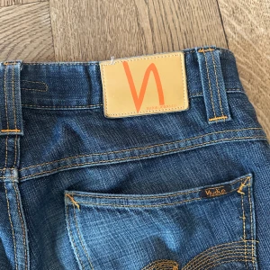Bootcut ola raw Nudie Jeans - Säljer ett par klassiska mörkblå jeans från Nudie Jeans med snygga kontrastsömmar och ikonisk logga på bakfickan. Jeansen har rak passform, fem fickor och är tillverkade i slitstarkt bomullsdenim. Perfekta för dig som gillar stilrena och tidlösa jeans.