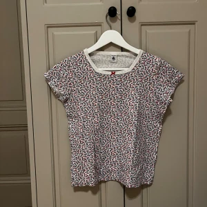 Vit blommig topp från Petit Bateau - Söt topp från Petit Bateau i vit med små blommor i rött och blått. Rund halsringning med vit kant och en liten röd rosett framtill. Kortärmad och tillverkad i mjuk bomull, perfekt för dig som gillar färgglada och lekfulla prints.