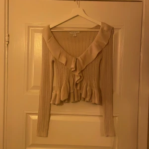 Beige stickad blus med volang från New Yorker - Är i storlek L men passar mig som har S/M. Helt ny utan prislap. Supersöt beige stickad blus från New Yorker med lång ärm, v-ringning och bred volang längs halsringningen och nederkanten. Blusen har dekorativa knappar framtill och ett ribbat, hålmönstrat tyg.