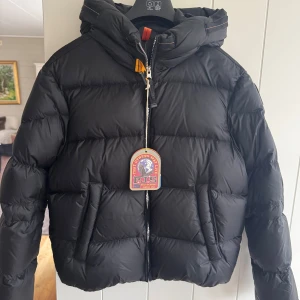 Ny- Svart pufferjacka från Parajumpers med tags , stl XL - Säljer en svart pufferjacka från Parajumpers med huva och coola detaljer som orange och gul rem vid halsen. Jackan har grova horisontella sömmar, två sidofickor och dragkedja framtill. Perfekt för dig som vill ha en varm och snygg jacka i vinter. Säljs på Kids Brandstore för 6995:-. Passar till stl 32-36