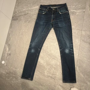 Mörkblå  jeans från Nudie - Snygga mörkblå skinny jeans från Nudie med klassiska fem fickor och orangea sömmar. Jeansen har slitningar och tvättade detaljer på knän och lår för en cool, använd look. Perfekta för dig som gillar en smal passform och stilren design.