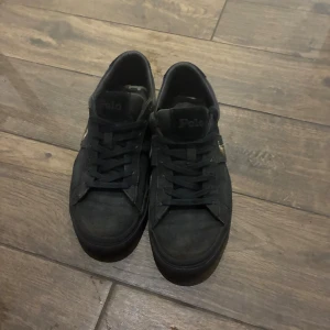 Svarta sneakers från Polo Ralph Lauren - Svarta sneakers från Polo Ralph Lauren med klassisk låg profil och snörning. Skorna har en diskret logga på sidan och bruna gummisulor med Polo-tryck undertill. Ovandelen är i textil och detaljerna är ton-i-ton för en clean look.