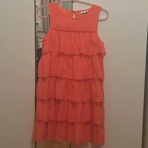 Korallfärgad volangklänning - Snygg korallfärgad kortklänning från C med flera lager volanger som ger en cool och lekfull look. Klänningen är ärmlös och har rund halsringning med en liten knapp i nacken. Perfekt för dig som gillar färg och volym.