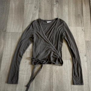 Omlott topp - Säljer en stilren grå/grön omlottopp från Nelly i mjuk bomull. Toppen har långa ärmar, v-ringad hals och knyt i midjan som ger en snygg siluett. Perfekt att styla med jeans eller kjol för en chill look. Fråga gärna om fler bilder eller info! Färgen heter ”taupe” på hemsidan!