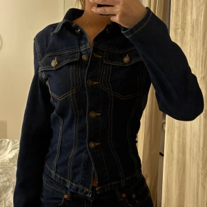 Mörkblå jeansjacka med knappar - Snygg mörkblå jeansjacka med klassiska guldfärgade sömmar och metallknappar framtill. Jackan har två bröstfickor med knapp och figurnära passform. Perfekt för dig som gillar en tidlös och cool look. Säljer även samma jeans storlek XXS, jeansjackan är storleken XS. Båda för 250kr, tröjan 170kr