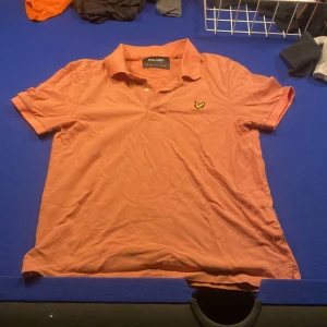 Rosa pikétröja från Lyle & Scott - Snygg rosa pikétröja från Lyle & Scott i storlek S. Tröjan har klassisk krage, korta ärmar och två knappar framtill. På bröstet sitter den ikoniska broderade gula fågeln. Materialet är mjuk bomull som andas och känns skönt mot huden.