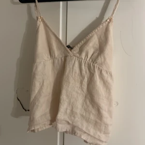 Brandy Melville topp - Säljer en ljusbeige linnetopp med smala justerbara axelband och v-ringad hals. Toppen har ett knytband i ryggen för extra detalj och passform. Tillverkad i mjukt linne, perfekt för varma dagar och chill stil.