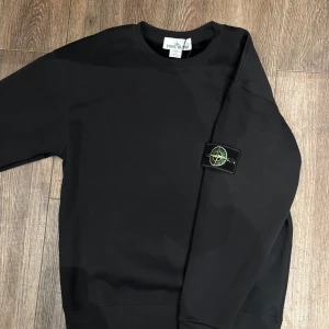Svart Stone Island Sweatshirt  - Svart sweatshirt från Stone Island med klassisk rund hals och ribbade muddar. På vänster ärm sitter den ikoniska Stone Island-patchen med broderad kompass i gult och grönt. Tröjan har en clean look och är gjord i mjuk bomull för skön komfort.