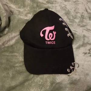 Svart TWICE keps med kedjor och ringar - Svart keps med rosa TWICE-logga framtill. Kepsen har coola detaljer som tre silvriga ringar på skärmen och fyra nitar med kedja på sidan. Justerbar baktill. Aldrig använd.