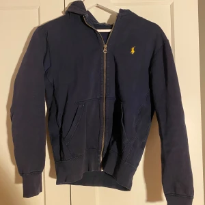 Ralph Lauren hoodie - Snygg marinblå hoodie från Ralph Lauren i storlek XS. Tröjan har huva, dragkedja framtill och två fickor. Klassisk gul logga broderad på bröstet. Mjuk bomullskvalitet och ribbade muddar vid ärmslut och nederkant.