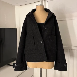 Svart kappa med stora knappar - Snygg svart kappa med klassisk krage och stora knappar framtill. Jackan har två fixkor och långa ärmar med knappdetalj vid mudden. Perfekt för dig som gillar en stilren och enkel look med lite oversized passform.