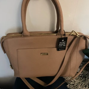 Beige rose handväska från Don Donna - Snygg beige rose handväska från Don Donna i stilren design med guldfärgade detaljer och dragkedja. Väskan har både handtag och axelrem, samt praktiska fack både på utsidan och insidan. Perfekt storlek för att få plats med allt du behöver.