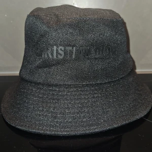 Svart bucket hat från Christian Dior - Stilren svart bucket hat med diskret logga framtill. Hatten har klassisk form med bredare brätte och är tillverkad i ett vävt tyg som ger en snygg struktur. Perfekt accessoar för att lyfta din outfit med exklusiv känsla.