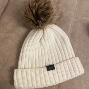 Vit ribbad mössa med pälsboll från Sätila - Snygg vit ribbstickad mössa från Sätila med stor brun pälsboll på toppen. Mössan har uppvikt kant och en liten svart logotyp framtill. Perfekt för kalla dagar när du vill vara både varm och stilren.