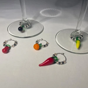 Glasmarkörer med färgglada grönsaker - Fem unika markörer med pärlor och hängande grönsaksberlocker i glas. Motiven är aubergine, jordnöt, chili, pumpa och majs. Perfekt för dig som gillar färg och förfina dina tillställningar.